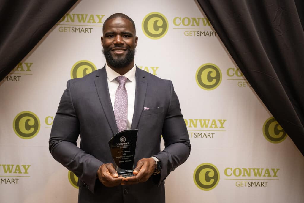 A young man in a suit holding an award in front of a Conway branded backdrop.