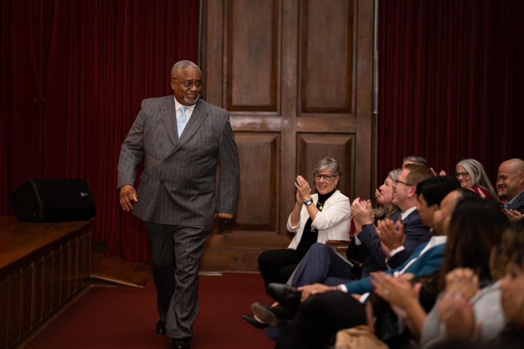 A man dressed in a suit walks down the front of the room while the audience applauds.