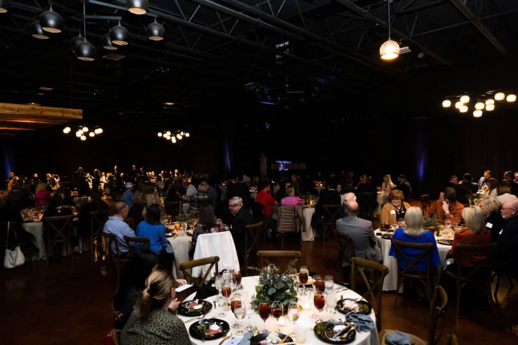 An event venue full of people sitting at round tables, celebrating a ribbon cutting event.
